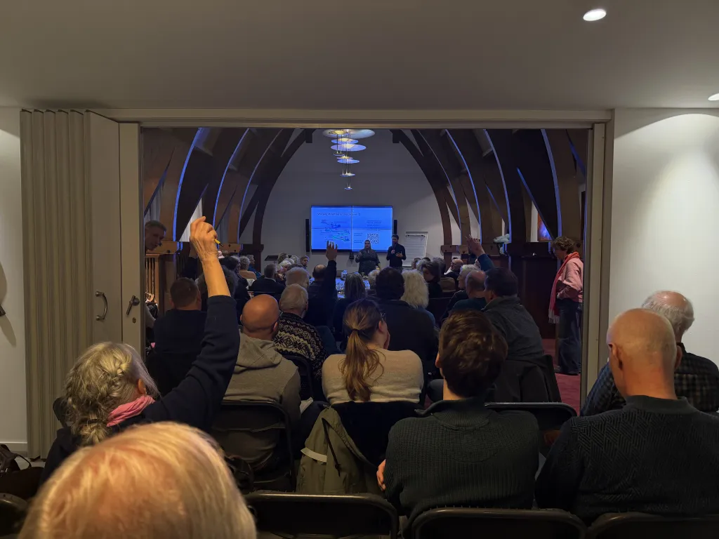 Een volle zaal met inwoners die zitten en luisteren naar een presentatie tijdens de informatiebijeenkomst voor VvE's. Voorin staat een spreker naast een groot scherm met een presentatie. Een persoon in het publiek steekt een hand op om een vraag te stellen.