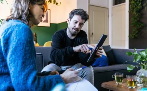 Een man en een vrouw zitten op de bank. De man heeft een iPad vast en geeft de vrouw uitleg over energiezuinig wonen.