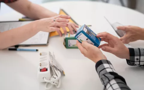 Je ziet twee paar handen die op een tafel energiebesparende producten pakken, zoals een ledlamp en een stekketblok met schakelaar 