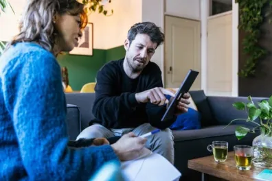 Een man en een vrouw zitten op de bank. De man heeft een iPad vast en geeft de vrouw uitleg over energiezuinig wonen.