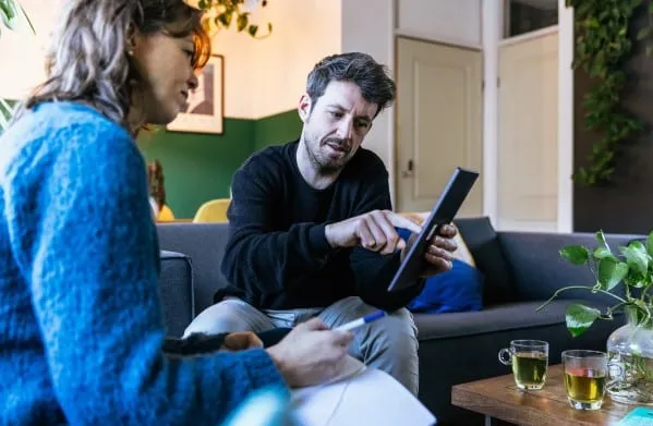 Een man en een vrouw zitten op de bank. De man heeft een iPad vast en geeft de vrouw uitleg over energiezuinig wonen.
