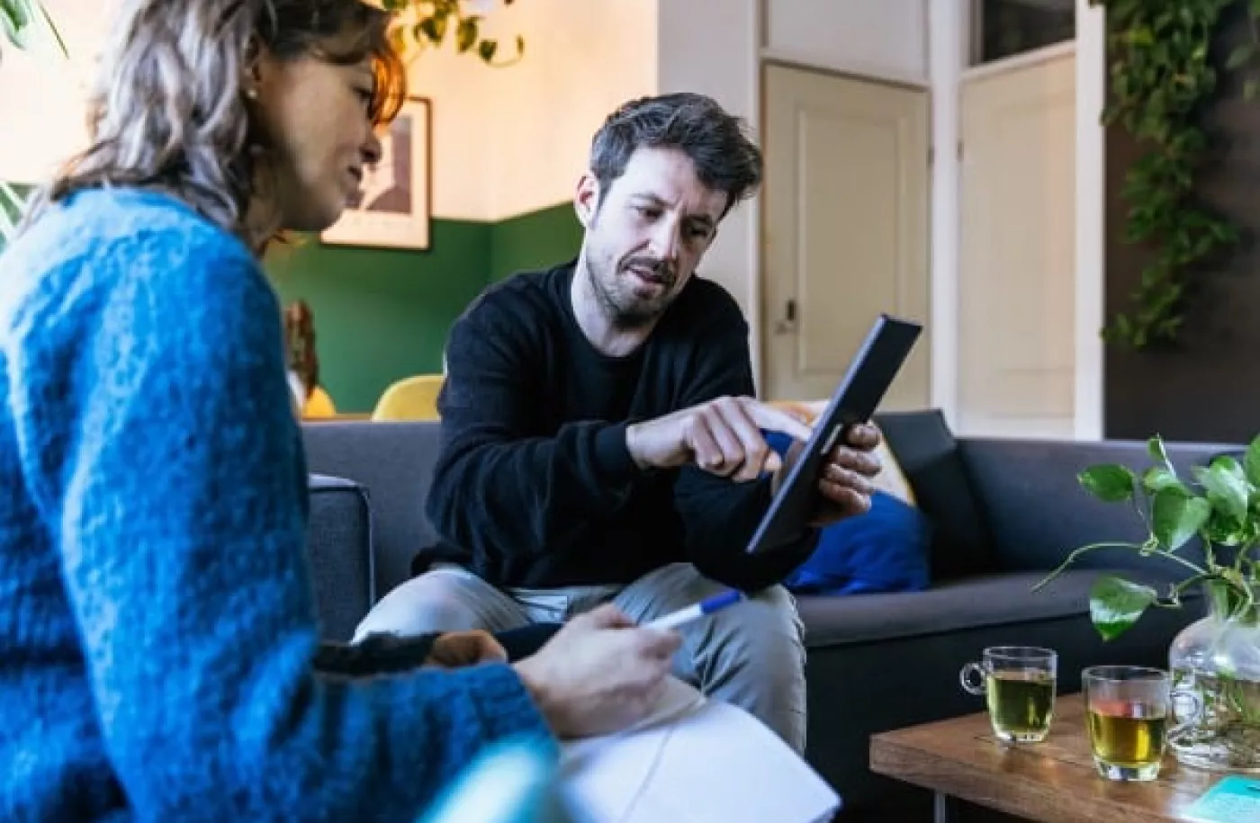 Een man en een vrouw zitten op de bank. De man heeft een iPad vast en geeft de vrouw uitleg over energiezuinig wonen.